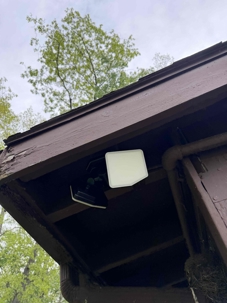 A white square light fixture is mounted under the eaves of a dark brown wooden building. Green trees are visible against a cloudy sky in the background.